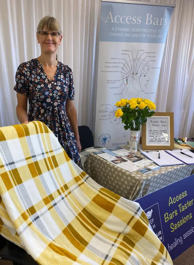 Renate standing beside her Access Bars stand at a wellbeing event, with yellow roses and a chart of the 32 points behind her